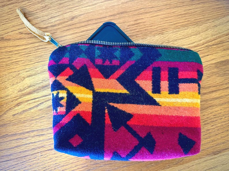 May include: A rectangular, zippered pouch with a colorful geometric pattern in shades of red, orange, yellow, green, and navy blue. The pouch has a leather wrist strap attached to the zipper.