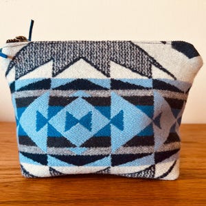 May include: A small, rectangular pouch with a geometric pattern in shades of blue, grey, and white. The pouch has a zip closure with a blue pull tab. The design features triangles and diamond shapes.