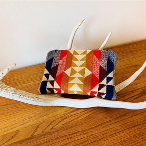 May include: A rectangular wallet with a geometric pattern in red, blue, yellow, and brown rests on a white antler. The wallet's design features triangles and other shapes. The antler is placed on a wooden surface.