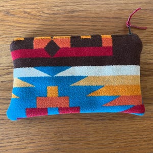 May include: A rectangular, colorful pouch with a zipper closure. The pouch features a geometric pattern in shades of brown, red, orange, blue, and white. A small, red leather pull is attached to the zipper.