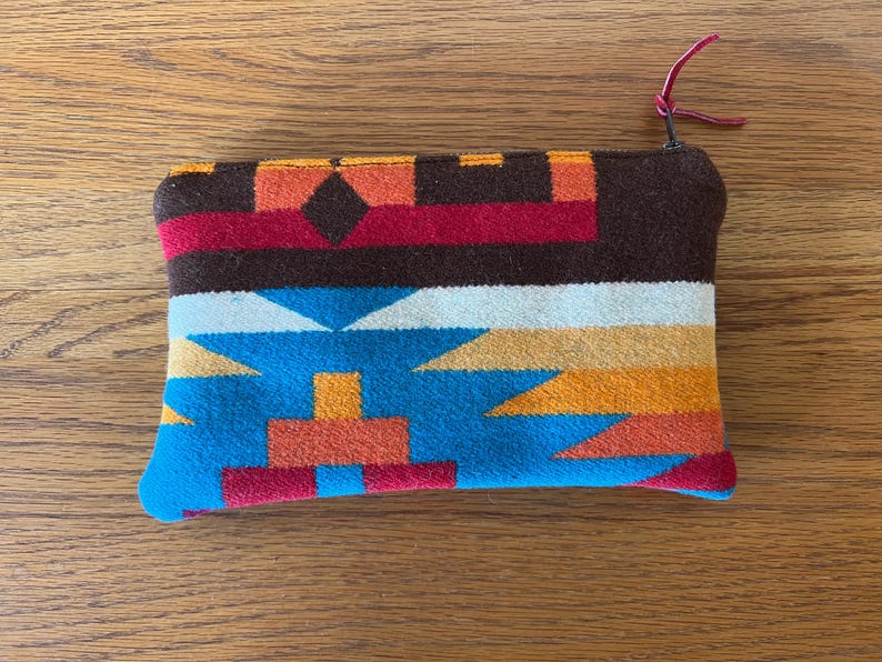 May include: A rectangular, colorful pouch with a zipper closure. The pouch features a geometric pattern in shades of brown, red, orange, blue, and white. A small, red leather pull is attached to the zipper.