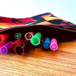 May include: A colorful pencil case with a geometric pattern, open to reveal a collection of colorful markers. The markers are various colors, including red, green, blue, and purple. The case is on a wooden surface.