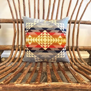 May include: A square decorative pillow with a geometric pattern in shades of red, orange, yellow, and blue. The pillow is set against a rustic wooden bench, creating a cozy, inviting scene.