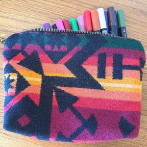 May include: A zippered pouch with a colorful geometric pattern in shades of red, orange, and black. The pouch is filled with various colored markers. A leather strap is attached to the zipper.