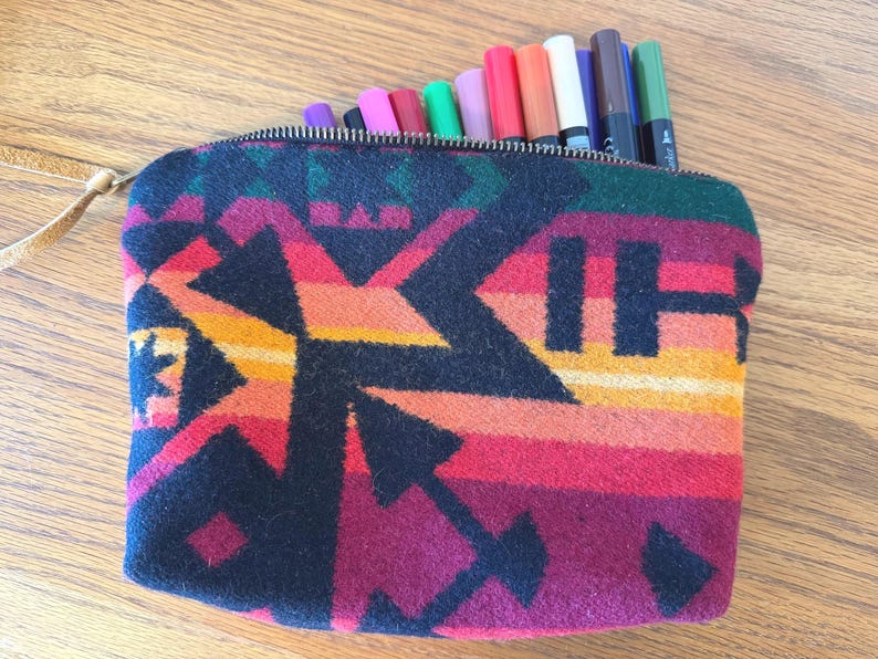 May include: A zippered pouch with a colorful geometric pattern in shades of red, orange, and black. The pouch is filled with various colored markers. A leather strap is attached to the zipper.