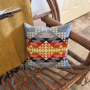May include: A square throw pillow featuring a geometric pattern in shades of light blue, dark blue, red, orange, and yellow. The design consists of repeating triangles and diamond shapes arranged in a symmetrical, centered composition on the fabric.