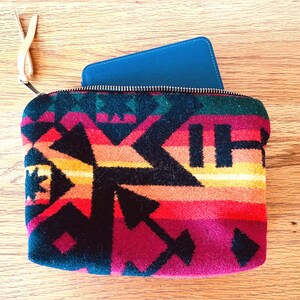 May include: A colorful zippered pouch with a geometric pattern in shades of red, orange, yellow, and black. A blue item is partially visible inside the pouch. The pouch has a leather pull tab.