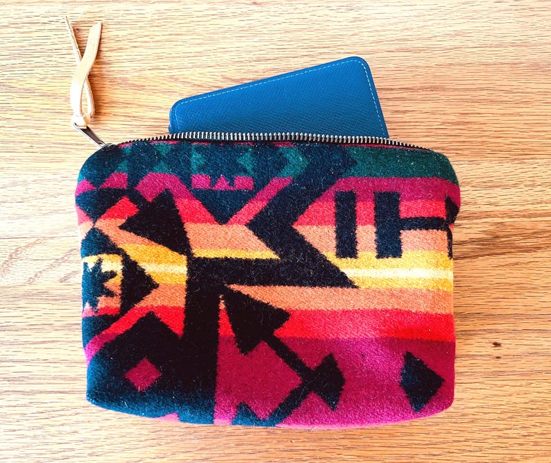 May include: A colorful zippered pouch with a geometric pattern in shades of red, orange, yellow, and black. A blue item is partially visible inside the pouch. The pouch has a leather pull tab.