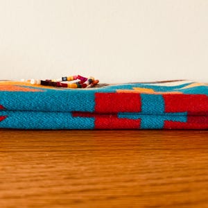 May include: A rectangular, folded textile item with a vibrant geometric pattern in turquoise, red, orange, and white. The item is adorned with a beaded embellishment. The item is resting on a wooden surface.