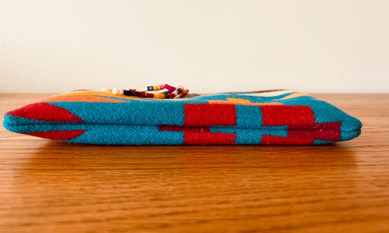 May include: A rectangular, folded textile item with a vibrant geometric pattern in turquoise, red, orange, and white. The item is adorned with a beaded embellishment. The item is resting on a wooden surface.