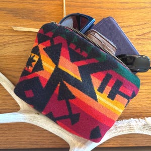 May include: A colorful zippered pouch with a geometric pattern in red, orange, yellow, and black. The pouch contains sunglasses, a wallet, and a small brown case. The pouch rests on a white antler against a wooden background.