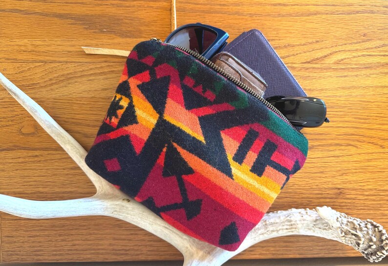 May include: A colorful zippered pouch with a geometric pattern in red, orange, yellow, and black. The pouch contains sunglasses, a wallet, and a small brown case. The pouch rests on a white antler against a wooden background.