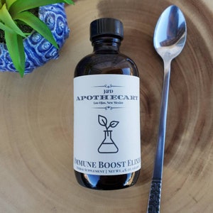 May include: A brown glass bottle with a white label that reads "J&D APOTHECARY Los Ojitos, New Mexico" and "IMMUNE BOOST ELIXIR HERBAL SUPPLEMENT | NET WT. 4 OZ (118 ml)". The bottle is sitting on a wooden surface with a silver spoon.