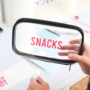 May include: A clear plastic zippered pouch with a black zipper and black trim. The pouch has the word "SNACKS" printed in pink letters.