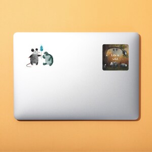 May include: A laptop sticker with a cartoon opossum and turtle holding a flower. The sticker says "Life is wild."