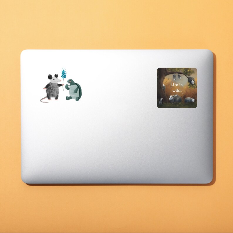 May include: A laptop sticker with a cartoon opossum and turtle holding a flower. The sticker says "Life is wild."