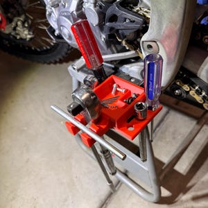 May include: A red plastic tool tray, branded with "HONDA", holding various tools, including screwdrivers, sockets, and a ratchet wrench. The tray is positioned near a motorcycle engine.