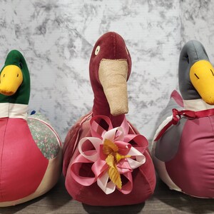 May include: Three plush toy ducks, one green, one grey, and one red, with pink and white bows. The ducks are sitting on a white textured surface.