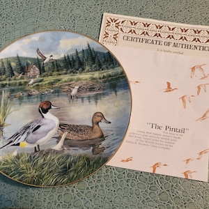 May include: A porcelain plate featuring a painting of a pintail duck in a lake setting. The plate is part of a collection called "Living with Nature: First Issue in the Collector's Plate Series by wildlife artist Ron Jenner." The plate is certified authentic by the Edwin M. Knowles China Company.