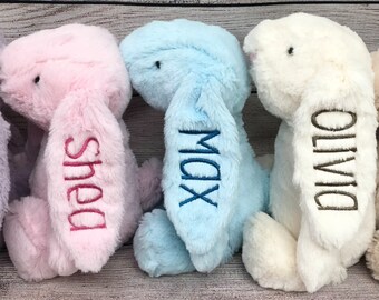 May include: Four plush bunny toys in different colors, each with a name embroidered on the ear. The bunnies are pink, blue, white, and beige. The names are Shea, Max, Olivia, and a fourth name that is not visible.