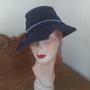 May include: A navy blue hat with a wide brim and a light gray band. The hat is displayed on a mannequin head with red lipstick. The hat's design includes a pleated section around the crown.