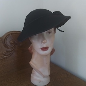 May include: A black hat with a wide brim and a decorative bow. The hat is displayed on a mannequin head with red lipstick. The hat is a vintage style, likely from the mid-20th century.
