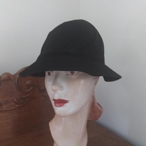 May include: A black cloche hat with a slightly curved brim. The hat is made of a soft material and has a textured crown. The hat is displayed on a mannequin head with red lipstick.