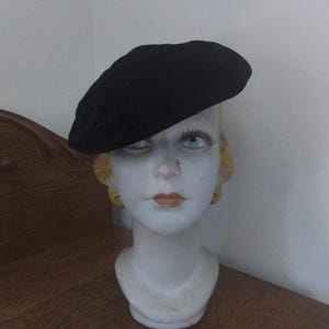 May include: A black velvet beret sits atop a mannequin head. The beret is a classic style, with a rounded crown and a close fit. The mannequin head is painted white with blue eyes and orange lips.