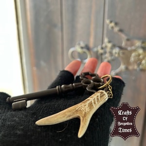 Peut inclure: Une clé vintage avec un charme d'antler décoratif attaché à une chaîne. La clé et le charme sont tenus dans une main gantée. L'arrière-plan est une porte en bois avec des ouvrages en métal complexes. L'image comprend le texte "Crafts of Forgotten Times".