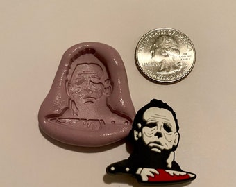 May include: A silicone mold for making a Halloween-themed cake topper. The mold is shaped like a person's head with a white mask and a knife. The mold is next to a black and white cake topper of the same design.