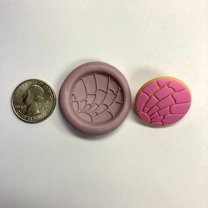 May include: A pink and brown concha cookie mold and a pink and brown concha cookie. The mold is made of silicone and has a detailed design. The cookie is about the size of a quarter.