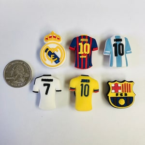 May include: Seven shoe charms featuring soccer team logos and jerseys. The charms include a white jersey with the number 7, a yellow jersey with the number 10 and the name "JAMES", a blue jersey with the number 10, a red and blue jersey with the number 10 and the name "MESSI", a white and gold logo with a crown, and a red and blue logo with the letters "FCB".