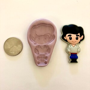 Pu&ograve; includere: Uno stampo in silicone viola di un principe dei cartoni animati con una camicia bianca e pantaloni blu. Lo stampo &egrave; accanto a una piccola statuina di plastica dello stesso principe dei cartoni animati.
