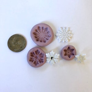 May include: Five silicone snowflake molds, two are purple and three are white. The molds are for making fondant or polymer clay snowflakes.