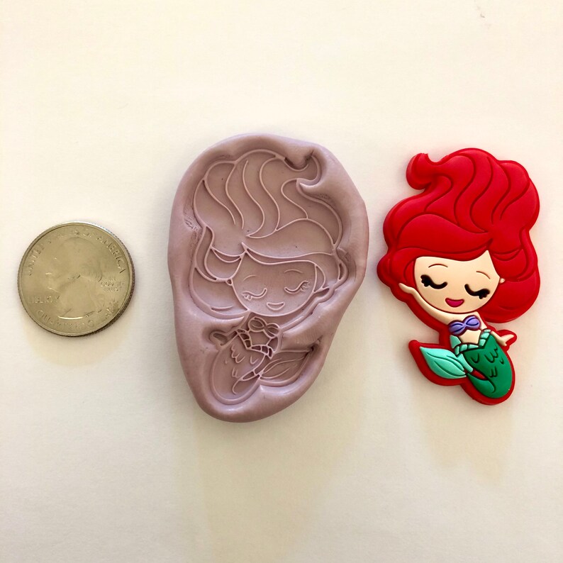 Pu&ograve; includere: Uno stampo in silicone e una statuetta di sirena rossa, verde e viola. La sirena ha lunghi capelli rossi, occhi chiusi e un reggiseno a conchiglia viola. Lo stampo &egrave; viola e mostra il contorno della sirena.