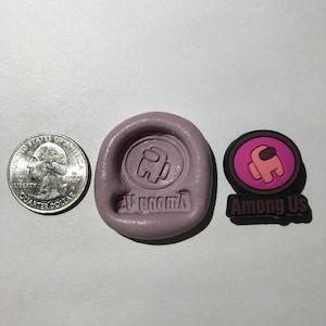 May include: A silicone mold for making Among Us character cake toppers. The mold is purple and has the Among Us character in the center. The mold is next to a small pink and brown Among Us character cake topper with the words "Among Us" printed on the bottom.