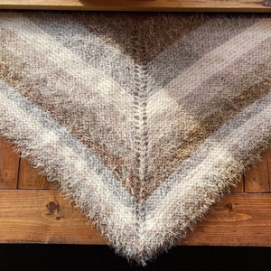 May include: A triangular shawl with a fuzzy texture in shades of brown, white, and gray. The shawl is made of yarn and has a fringed edge.