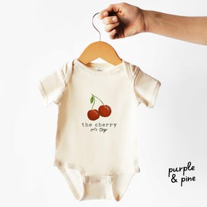 May include: A cream-colored baby bodysuit featuring a graphic of two red cherries with a green leaf. Below the cherries, the text reads "the cherry on top" in a black typewriter-style font. The garment is displayed on a wooden hanger.