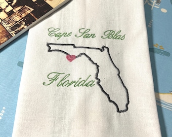 May include: White embroidered towel with the outline of Florida and a heart marking a location. The words "Cape San Blas" and "Florida" are embroidered in green script. A book titled "Gulf County" is in the background.