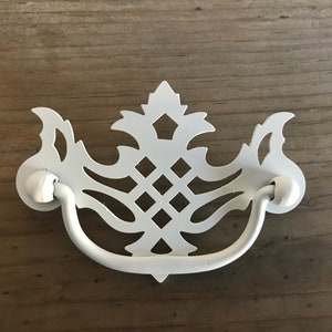 May include: White metal cabinet pull with a decorative, ornate design. The pull has a curved handle and a center design with a diamond pattern.