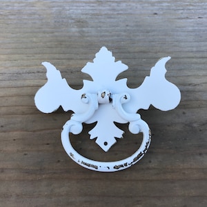 Könnte beinhalten: Weißer Metall-Schubladengriff mit einem dekorativen, verzierten Design. Der Griff hat einen Ringgriff und ein Blumenmuster.