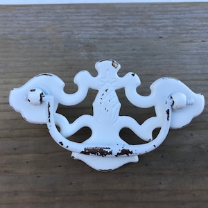 Könnte beinhalten: Weißer Metall-Schubladengriff mit einem dekorativen, verzierten Design. Der Griff hat eine abgenutzte Oberfläche und einen Schleifen-Griff.