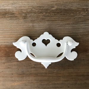May include: White metal drawer pull with a heart-shaped cutout and ornate details. The pull has a curved handle and is designed for use on cabinets or drawers.