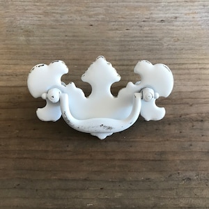 May include: White metal drawer pull with a decorative, ornate design. The pull has a distressed finish and a slightly aged look.