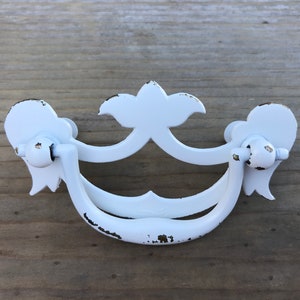 Puede incluir: Tirador de armario de metal blanco con acabado desgastado y diseño ornamentado. El tirador tiene un asa curva y dos pomos decorativos a los lados.