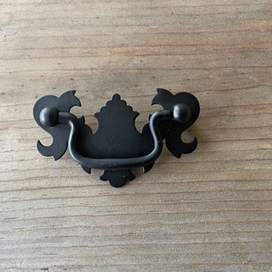 May include: Black metal drawer pull with a decorative, ornate design. The pull has a curved handle and two decorative knobs on either side.