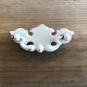 May include: White distressed drawer pull with a decorative design. The hardware features a central oval plate with ornate detailing and curved handles on each side. The pull is painted white with visible wear and tear, suggesting a vintage aesthetic.