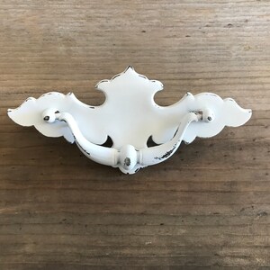 Op de afbeelding: Witte metalen ladetrekker met een decoratief, sierlijk ontwerp. De trekker heeft een verweerde afwerking, waardoor hij een vintage uitstraling heeft.