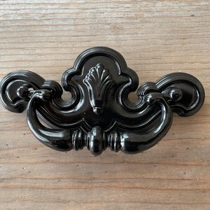 May include: Black metal cabinet pull with a decorative, ornate design. The pull has a curved shape and a center detail that resembles a flower.
