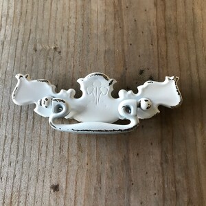 May include: White metal drawer pull with a decorative design and a curved handle. The pull has a distressed finish.